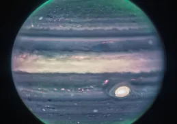 Jupiter dominates the black background of space. The image is a composite, and shows Jupiter in enhanced color. The planet’s Great Red Spot appears white here. The planet is striated with swirling horizontal stripes of neon turquoise, periwinkle, light pink, and cream. The stripes interact and mix at their edges like cream in coffee. Along both of the poles, the planet glows in turquoise. Bright orange auroras glow just above the planet’s surface at both poles.
