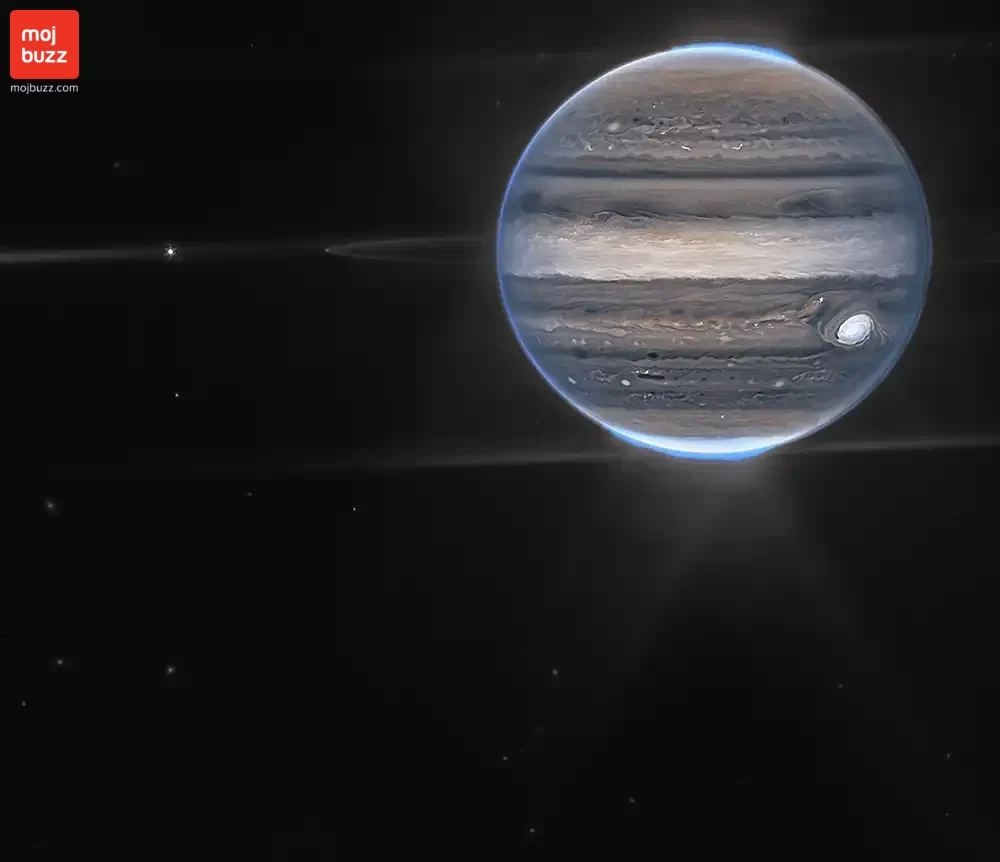 A wide field view showcases Jupiter in the upper right quadrant. The planet’s swirling horizontal stripes are rendered in blues, browns, and cream. Electric blue auroras glow above Jupiter’s north and south poles. A white glow emanates out from the auroras. Along the planet’s equator, rings glow in a faint white. These rings are one million times fainter than the planet itself! At the far left edge of the rings, a moon appears as a tiny white dot. This moon is only about 12 miles (20 km) across. Slightly further to the left, another moon, about 100 miles (150 km) across, glows with tiny white diffraction spikes. The rest of the image is the blackness of space, with faintly glowing white galaxies in the distance.